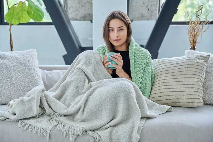 ソファにカップを手にした暖かい毛布の下に座っている美しい若い女性